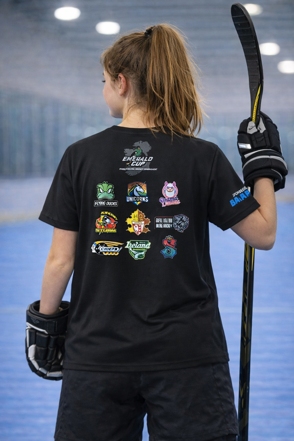 Person wearing a black t-shirt with logos and holding a hockey stick on an indoor ice rink.