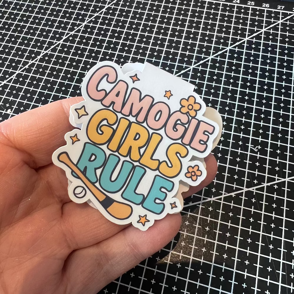 Hand holding a 'Camogie Girls Rule' sticker on a grid background