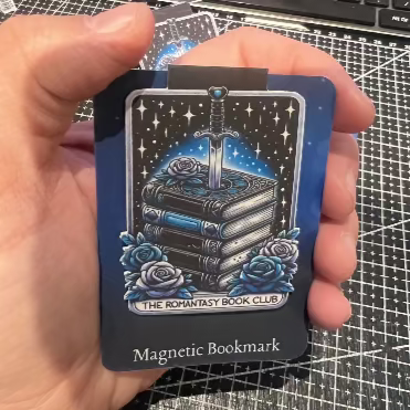 Hand holding a magnetic bookmark with a blue and black design on a grid-patterned surface.