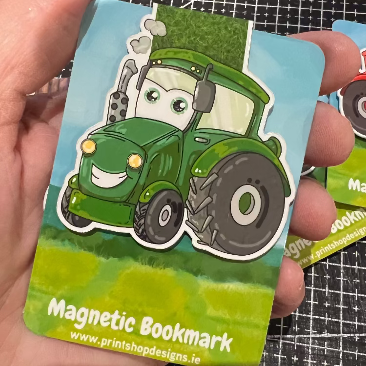 Magnetic bookmark with cartoon tractor design held by a hand on a black grid background