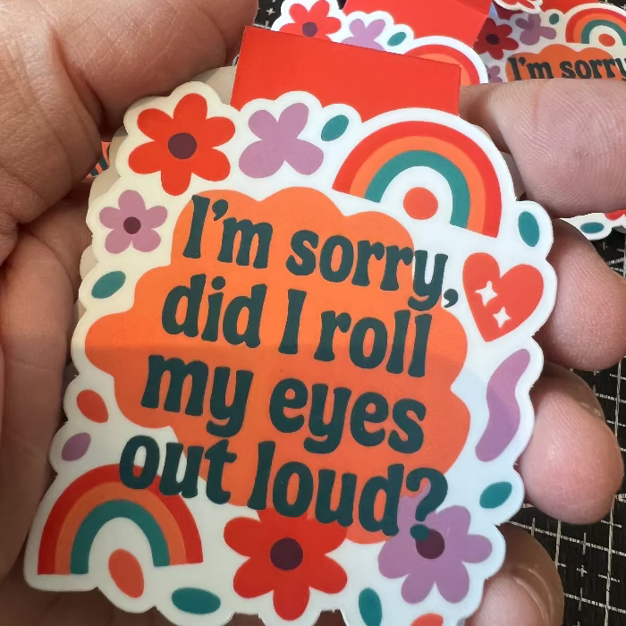Hand holding a colorful sticker with text 'I'm sorry, did I roll my eyes out loud?' on a checkered background.