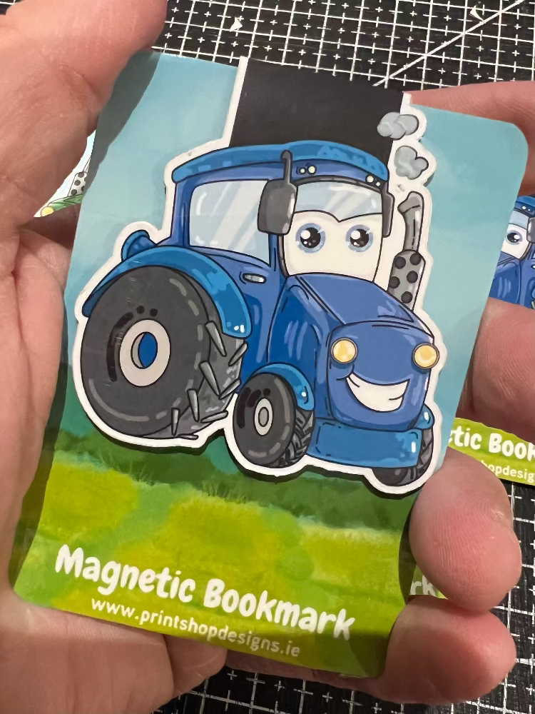 Magnetic bookmark with a cartoon blue tractor held by a hand on a grid background