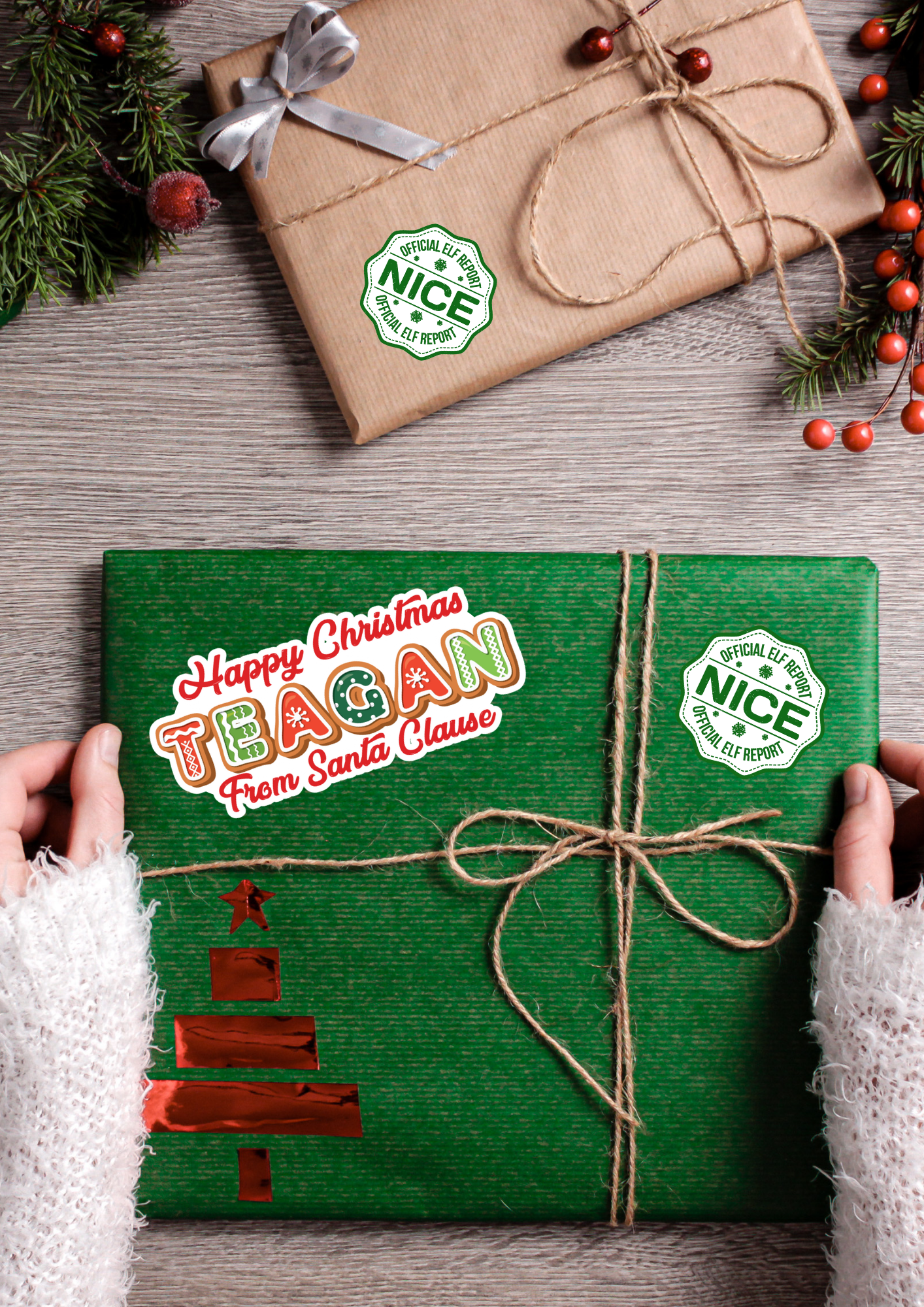 Green gift box with 'Happy Christmas Teagan from Santa Clause' text, tied with twine, on a wooden surface with a wrapped brown gift and Christmas decorations.
