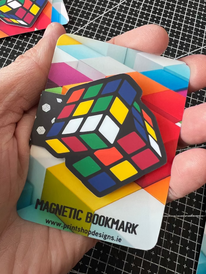 Colorful Rubik's Cube magnetic bookmark held by a hand on a black grid background.