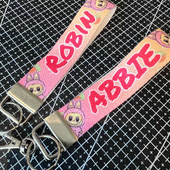 Two pink keychains with cartoon characters and names 'Robin' and 'Abbie' on a black grid background.